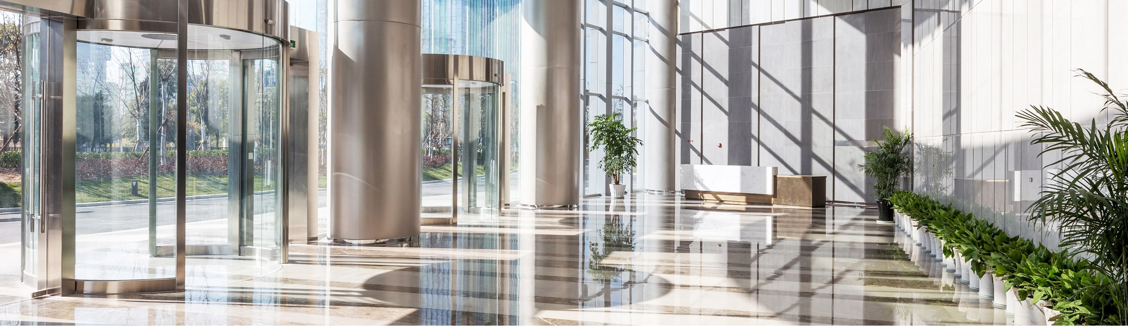 Modern office building lobby with tall glass windows, revolving doors, a marble reception desk, and rows of green potted plants along the walls.