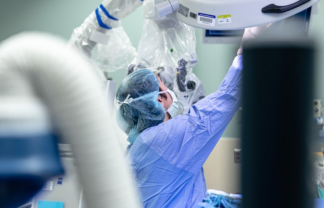 A medical professional in surgical attire adjusts equipment in an operating room, surrounded by medical devices and sterile tools.