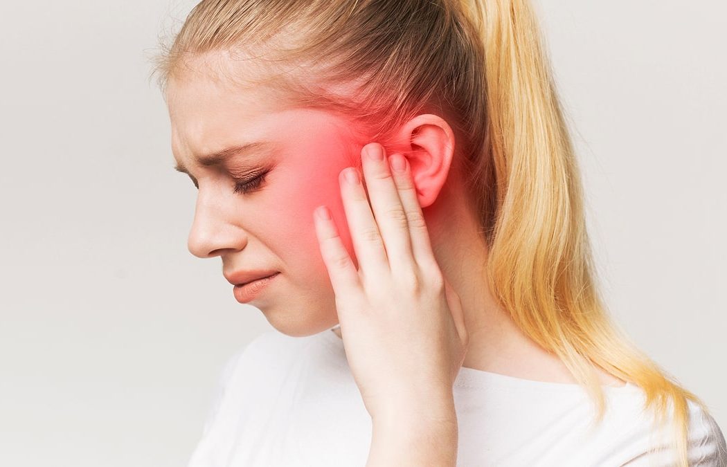 Young woman with long blonde hair holding her left ear, face contorted in pain, with a red area highlighting discomfort around her ear.