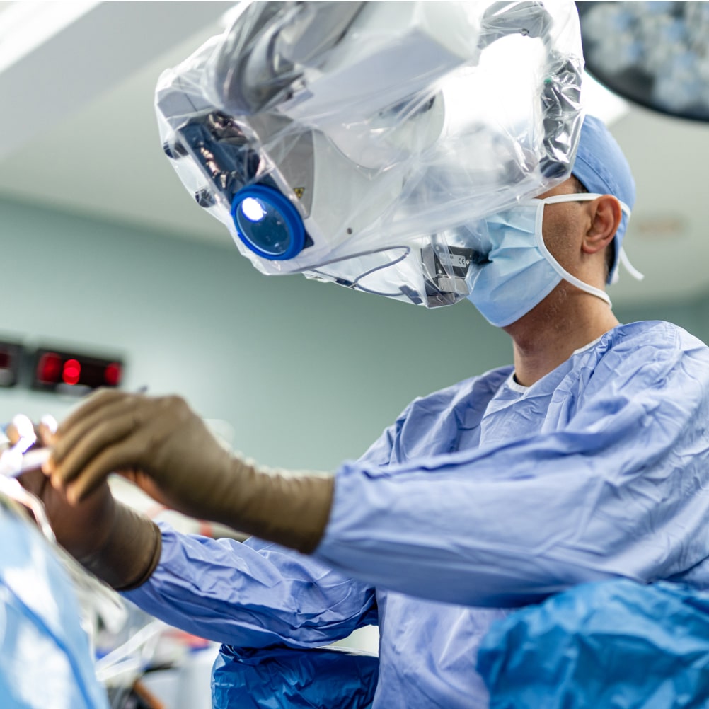 A surgeon wearing scrubs, gloves, and a mask operates while using a surgical microscope in a brightly lit operating room.