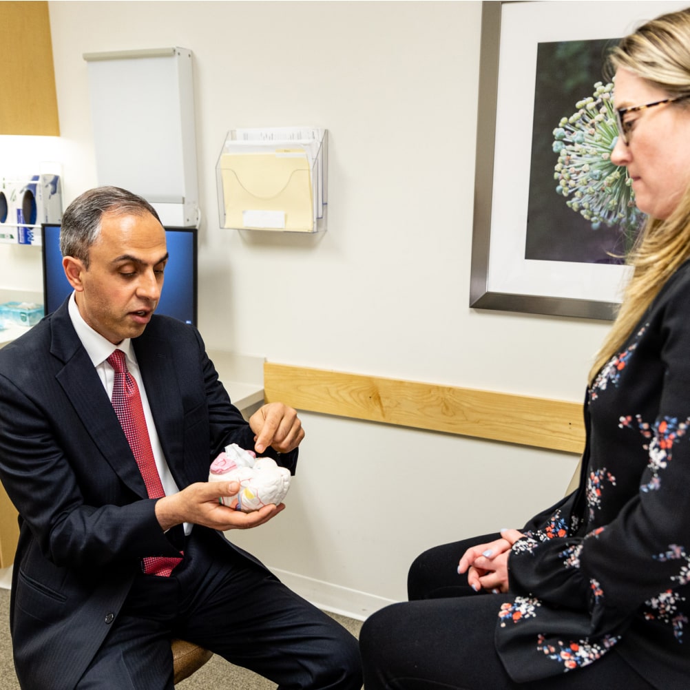 A doctor in a suit explains a medical model to a seated woman in an examination room.