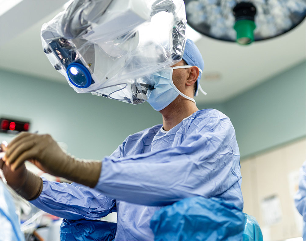 A surgeon in scrubs and a mask operates using a surgical microscope in an operating room.