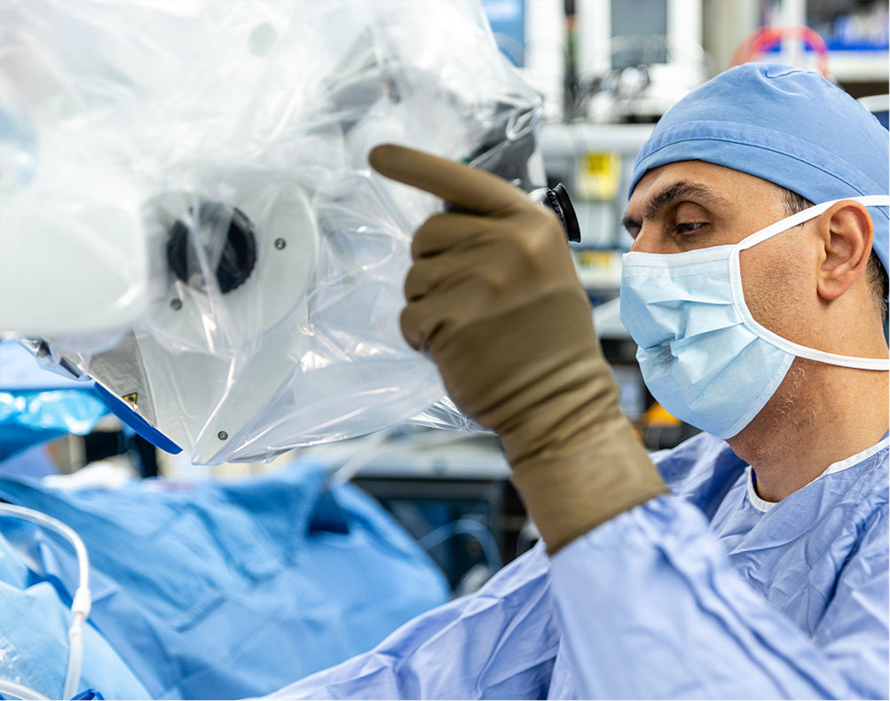A surgeon wearing scrubs, gloves, and a mask operates a medical device, possibly a microscope, during a surgical procedure.