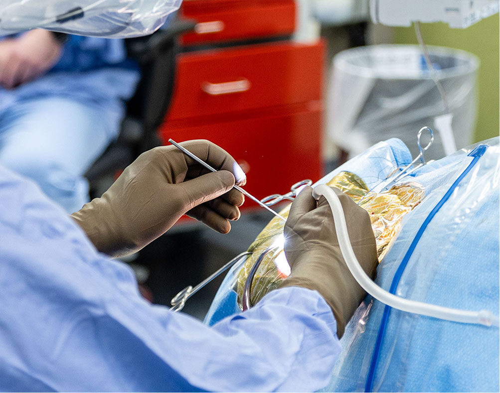 A medical professional wearing gloves and scrubs performs a surgical procedure, using tools and working on a patient covered with a sterile drape.