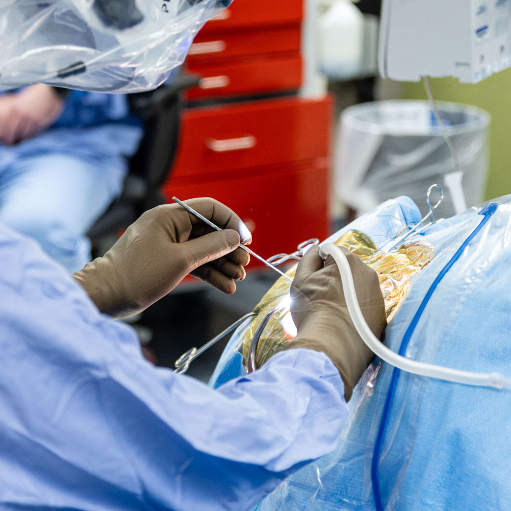 A surgeon wearing gloves and a sterile gown performs a medical procedure on a patient, using surgical instruments and tubing under bright light in an operating room.
