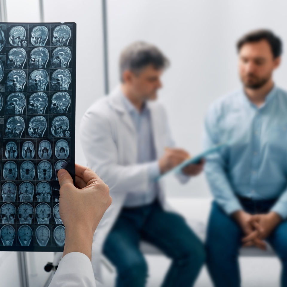 A doctor reviews brain scan images while another doctor speaks with a seated male patient in a clinical setting.